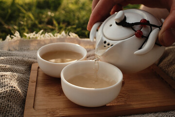 Hand pouring hot tea from a white teapot into a small porcelain cup outdoors on a wooden tray. Picnic in Chinese style for two people outdoor