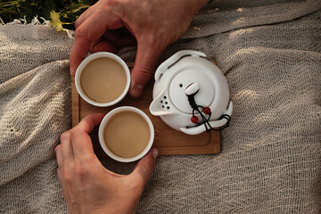 Hands holding two cups of tea beside a white teapot on a wooden tray placed on fabric outdoors. Top view. Picnic in Chinese style for two people outdoor