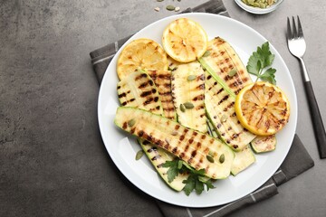 Delicious grilled courgette slices with pumpkin seeds, lemon and parsley served on grey textured table, flat lay. Space for text