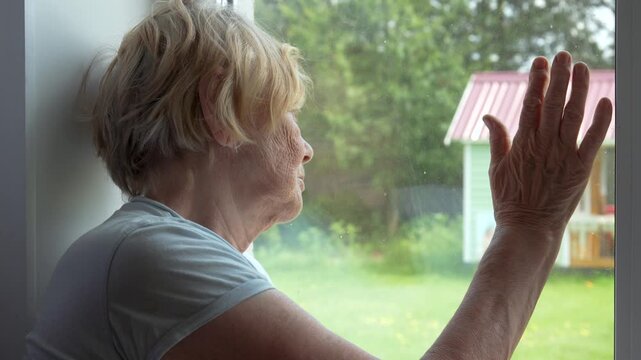 Elderly woman with gray hair and deep wrinkles on skin looks longingly out window at backyard. Hand touches glass, as if it is trying to reach outside world. Self isolation old ill people