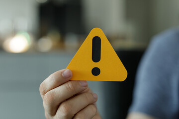Close-up of a hand holding a yellow triangle with an exclamation mark, signifying warning or caution.