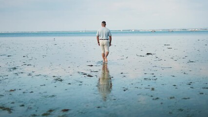 A man is walking on a beach with the ocean in the background. The man is wearing a white shirt and khaki shorts. The beach is covered in seaweed and the water is calm - Powered by Adobe