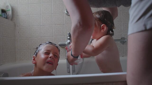 Mother helping wash older child&rsquo;s hair while toddler plays with cup during bath time, intimate family bonding moment, playful and cozy atmosphere in home bathroom