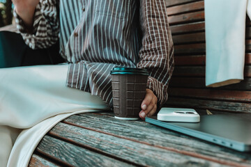 Close-up of coffee cup, smartphone, and laptop on wooden bench with woman's hand holding the cup, cozy work and lifestyle moment outdoors