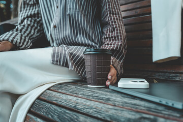 Fototapeta premium Close-up of coffee cup, smartphone, and laptop on wooden bench with woman’s hand holding the cup, cozy work and lifestyle moment outdoors