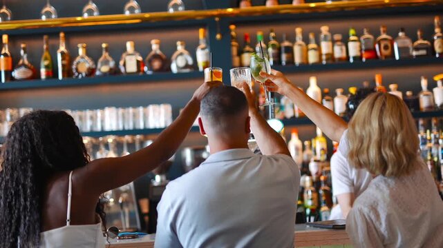 A group of people are celebrating with drinks at a bar. There are many bottles on the shelves behind them, and the people are raising their glasses in a toast. Scene is festive and social