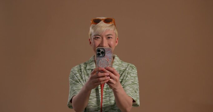 Video clip of a young adult male looking at the camera while taking photos on a mobile phone in the studio. He has bleached hair and is wearing casual clothing.