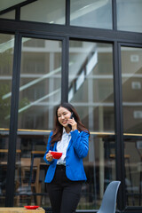 Fototapeta premium Asian businesswoman in suit using mobile phone while standing in front of modern business building.
