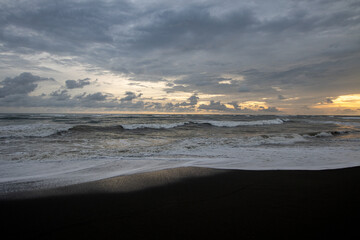 Dramatic seascape at sunset with dark sand beach and turbulent waves under cloudy sky