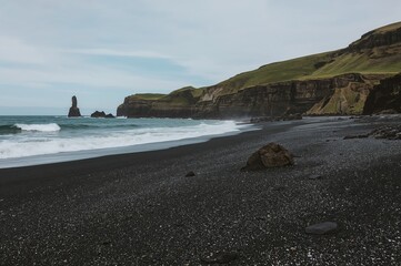 Dark sandy shore adjacent to a small coastal settlement with volcanic basalt columns and lush moss-covered hills by the ocean. Renowned travel location known for its unique natural features.