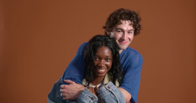 Medium video clip of two people embracing in the studio, the male standing behind his female friend. They are both smiling as they sway from side to side, wearing casual clothing.
