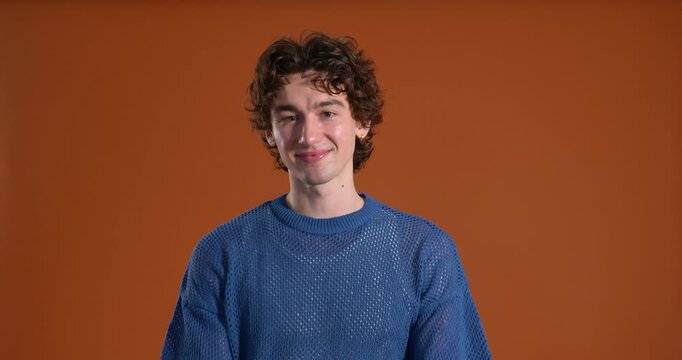 Medium video clip of a young adult male smiling while making peace signs towards the camera in the studio. He has dark curly hair and is wearing casual clothing.