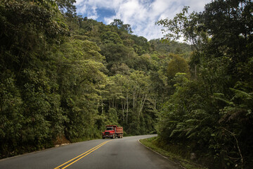 Red truck driving on a winding road through lush green forest landscape under a cloudy sky