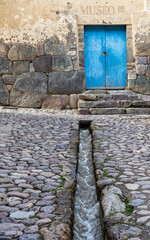 Narrow cobblestone street with a traditional water canal leading to a rustic blue door in Ollantaytambo Sacred Valley, Peru.