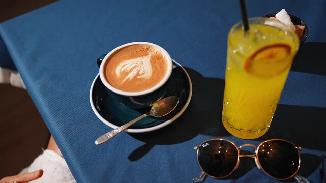 A cup of coffee and a glass of orange juice sit on a table. The coffee is in a blue mug with a white design, and the orange juice is in a glass with a straw. The tablecloth is blue