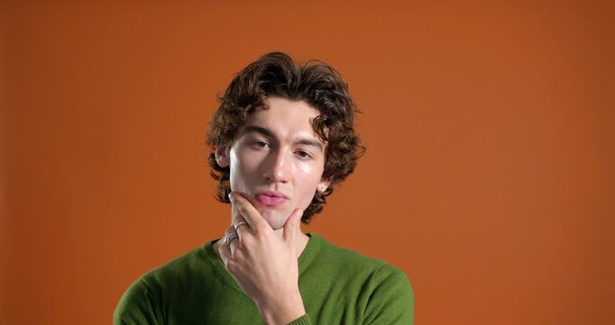  Medium video clip of a young adult male pondering with a serious expression in the studio, resting one hand on his chin thoughtfully. He has curly hair and is wearing casual clothing.