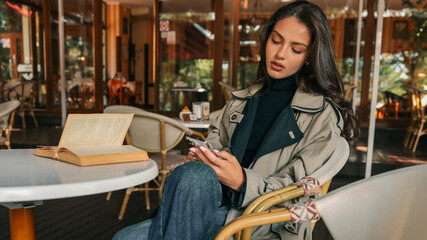 Young woman in trench coat sitting at outdoor cafe table with vintage book and smartphone. Autumn mood with elegant style and cozy ambience