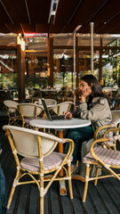 Side view of a young woman in a beige trench coat sitting at a white round table with a laptop in an outdoor cafй. She is focused and thoughtful, surrounded by empty chairs and soft natural light