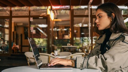 Confident young woman in a beige trench coat resting her head on hand while using a laptop at a white outdoor café table. Looking at camera with calm, thoughtful expression