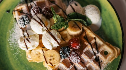 Tasty Belgian waffles served with banana slices, vanilla ice cream scoops, raspberries, blackberries, and chocolate syrup on a green ceramic plate dusted with powdered sugar