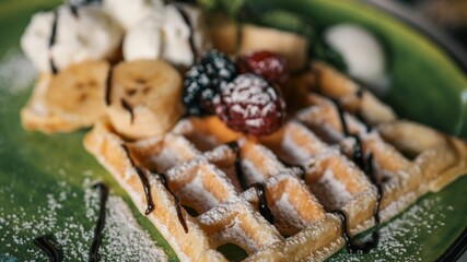 Tasty Belgian waffles served with banana slices, vanilla ice cream scoops, raspberries, blackberries, and chocolate syrup on a green ceramic plate dusted with powdered sugar
