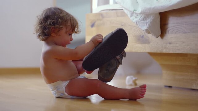 Toddler sitting on bedroom floor lifting sandal to examine sole closely, focused exploration, early childhood learning, curiosity, sensory development, family life