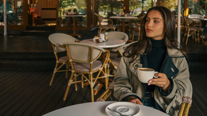 Young woman in a beige trench coat holding a cappuccino cup, seated at a round white table in a stylish cafй, looking thoughtfully into the distance