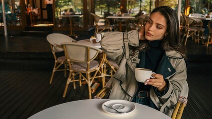 Young woman in a beige trench coat holding a cappuccino cup, seated at a round white table in a stylish café, looking thoughtfully into the distance
