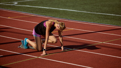 Female runner stands prepared at the start line on a track illuminated by sunlight with calm intensity. Concept of crouch start, focus, anticipation, mental strength, and explosive power before sprint