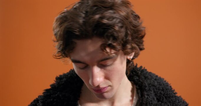 Close-up video clip of a young adult male posing for the camera in the studio, wearing gold novelty sunglasses and a black fur jacket. He has dark curly hair and is wearing a selection of rings.
