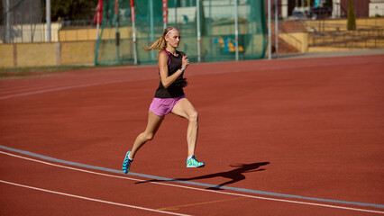 Fototapeta premium Young woman runs on empty track outside while training on sunny day with strength and focus. Concept of mid-run intensity, athletic balance, racing power, determination, and performance discipline.