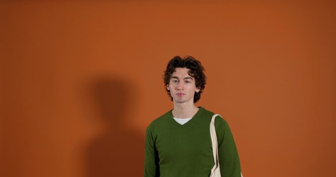 Medium video clip of a young adult male walking into frame, looking directly at the camera with a serious expression, before walking off-frame. He has curly hair and is wearing casual clothing