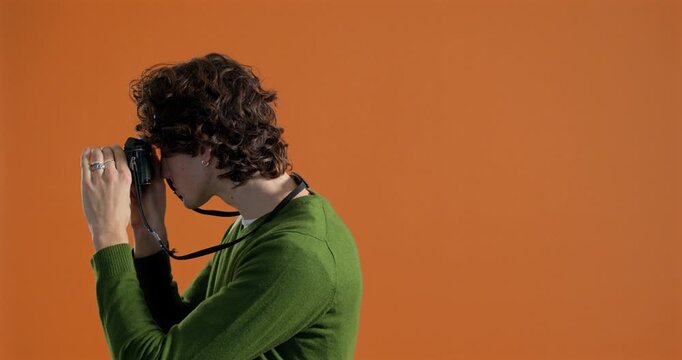 Medium video clip of a young adult male taking photograohs in the studio. He is facing one side before turning to face the camera. He has dark curly hair and is wearing casual clothing.
