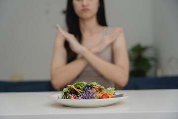 Young woman refusing to eat a plate of salad with hand gesture, showing dislike or aversion to vegetables. Concept of picky eating, diet frustration, food rejection, or emotional eating issues.