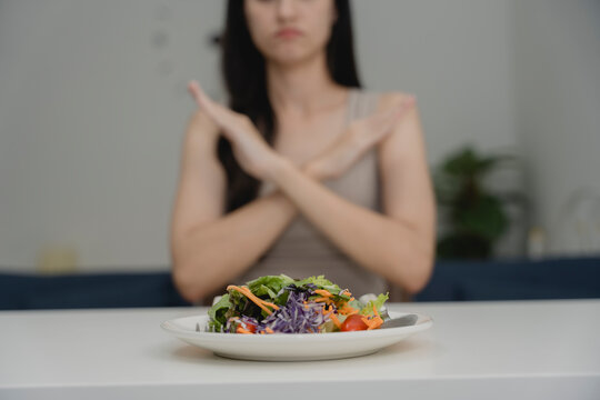Young woman refusing to eat a plate of salad with hand gesture, showing dislike or aversion to vegetables. Concept of picky eating, diet frustration, food rejection, or emotional eating issues. - Powered by Adobe