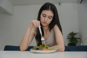 Young woman looking bored or unhappy while eating salad, pushing food with fork at table. Concept...