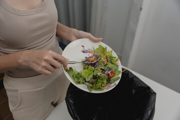 Woman scraping fresh salad into trash bin, representing food waste, poor appetite, sustainability issues, diet rejection, or environmental concerns about throwing away healthy meals.
