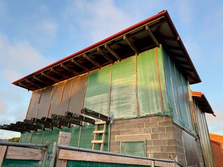 Unfinished building with green plastic sheeting and red metal roof captured at sunset. Wooden beams and cinder blocks show construction process and temporary weather protection.