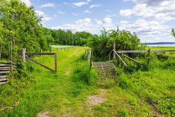 Wooden gate and rustic fence leading to green meadow with wooden footbridge and lake on the background. Sweden.