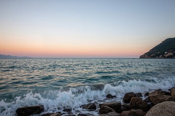 Obraz premium Tranquil seaside view at dawn with soft waves hitting rugged rocks, distant cliffs, and a tiny island on the edge of the sea. The sky displays gentle pastel shades of pink and blue.