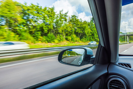 View from inside car with side mirror showing highway, traffic, bridge and roadside trees. Sweden.