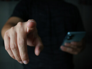  Close up of index finger pointing out with blurred background, other hand holding phone, dark tone and warm light hitting hand, business concept, checking goods, ordering via digital system.