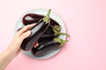 Woman holding fresh eggplant above plate with whole ones on pink background, top view. Space for text