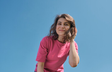 Beautiful carefree young woman with curly hair in pink t-shirt smiling and looking down at camera on blue sky background low angle shot. 