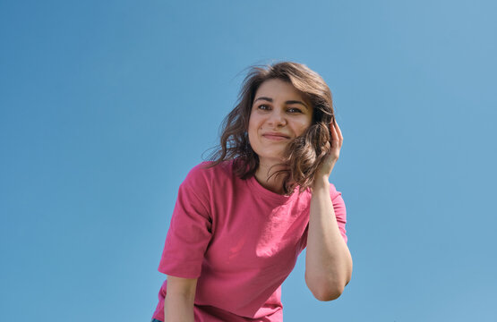 Beautiful carefree young woman with curly hair in pink t-shirt smiling and looking down at camera on blue sky background low angle shot. 