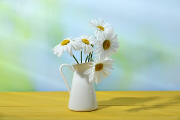 Beautiful chamomile flowers in vase on yellow wooden table against blurred background, closeup
