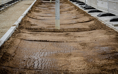 Obraz premium Urban Construction Site With Freshly Leveled Soil And Concrete Sidewalk Borders: Street Renovation Project With Safety Barriers And Building Background