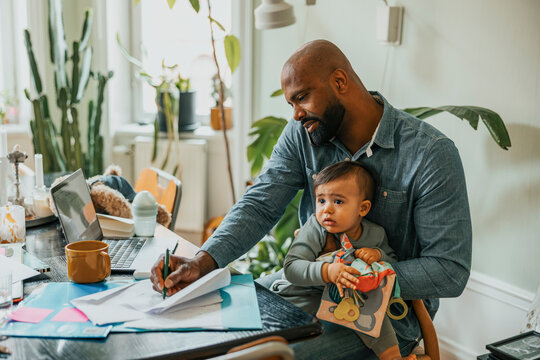 Businessman talking on smart phone and taking notes while sitting with son at home