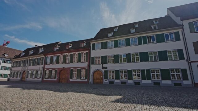 Wide shot of historic row houses with colorful green shutters on M&uuml;nsterplatz, across from Basel Minster, under a clear blue summer sky in Basel, Switzerland. Filmed on July 25, 2025.