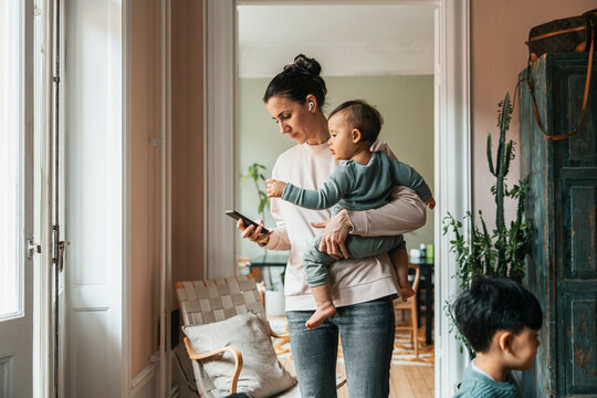 Woman wearing wireless in-ear headphones standing with baby boy while using smart phone at home